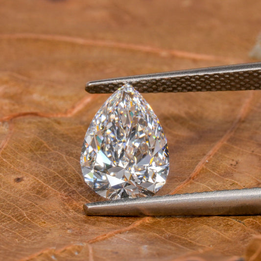 Loose pear-cut diamond, clear faceted gemstone held with tweezers on a leaf surface, close-up front view with pointed tip and rounded base