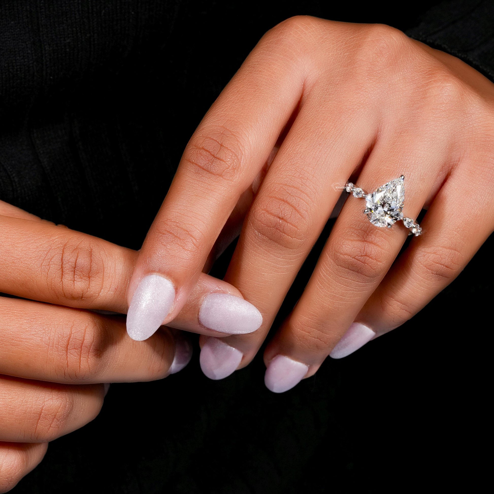 Engagement ring with polished white metal band, pear-shaped clear gemstone and round accent stones, worn on finger, angled view
