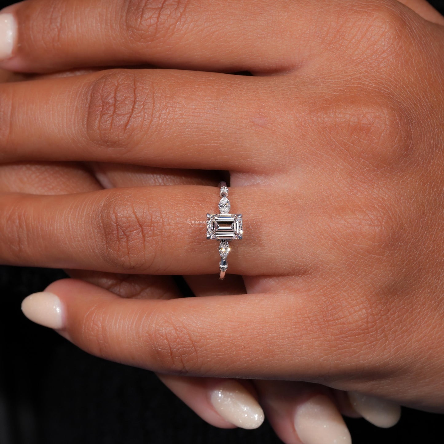 Engagement ring with white metal band, emerald-cut clear center stone, marquise side stones and round accents, worn on finger top view
