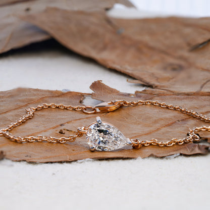 Bracelet with pear-shaped clear stone in yellow metal chain setting on dried leaf, angled close-up view, delicate chain bracelet jewelry
