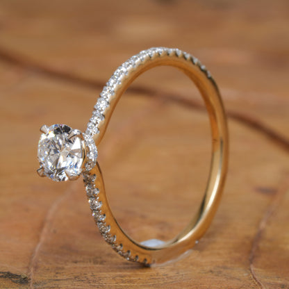 Engagement ring with round clear center stone, white metal pavé band, four-prong solitaire setting, angled side view on wooden surface
