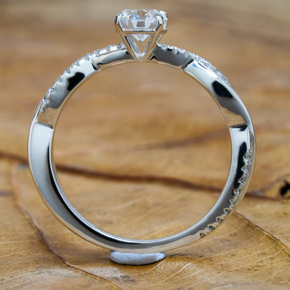 Ring with round clear solitaire, white metal twisted band with pavé-set shoulders, rear profile on wooden surface
