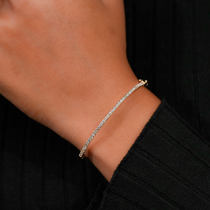 Bracelet with yellow metal band, slim round stone tennis design, worn on wrist at an angled front view against a black sleeve background
