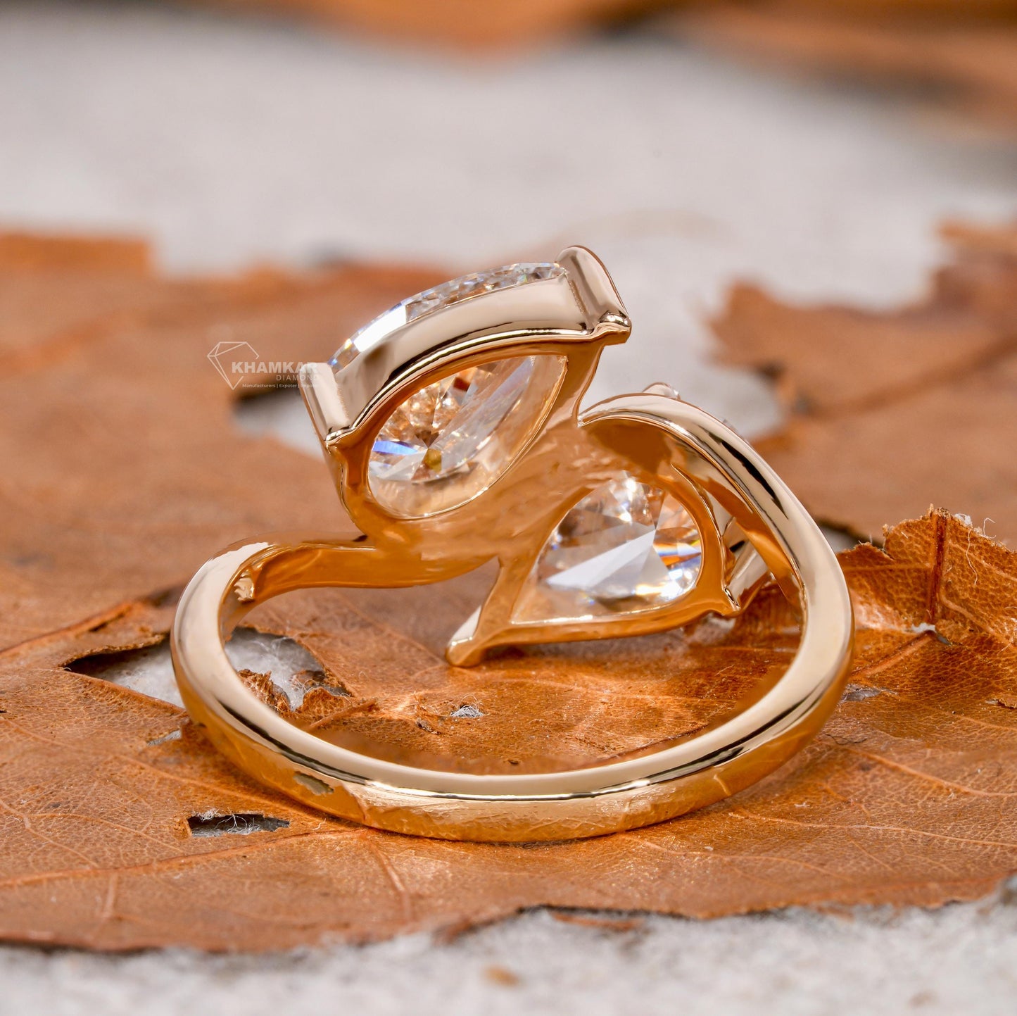 Engagement ring with yellow metal bypass band, two pear-shaped clear gemstones, open crossover setting, rear angled view on leaf background
