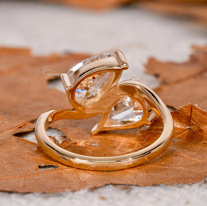 Engagement ring with yellow metal bypass band, two pear-shaped clear gemstones, open crossover setting, rear angled view on leaf background
