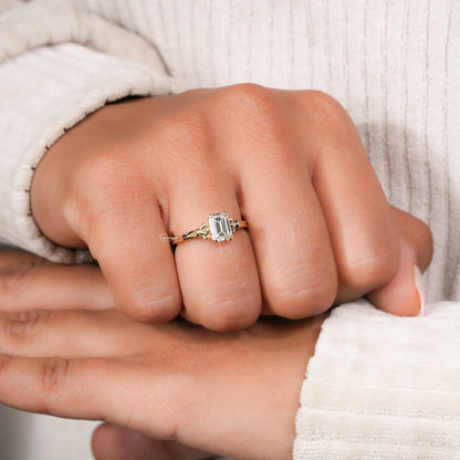 Engagement ring with rectangular clear gemstone, yellow metal band, halo design with round accent stones, worn on finger, angled view
