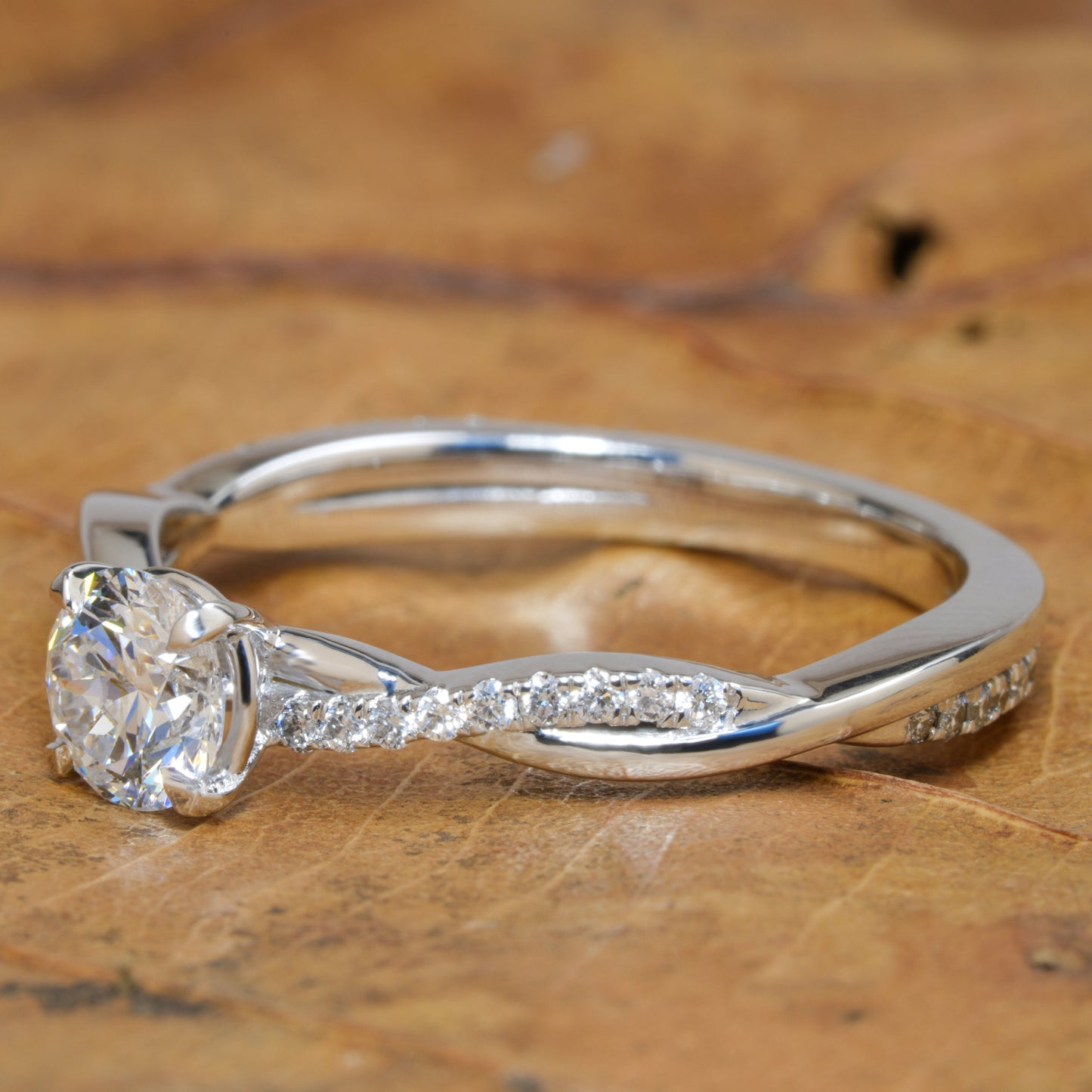 Ring with round clear center stone, twisted white metal band with pavé-set shoulders, side view on a wooden surface
