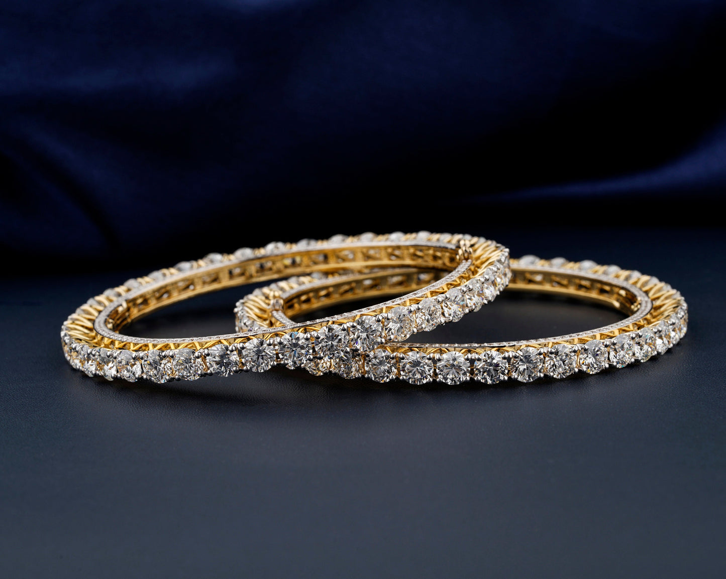 Bracelets with yellow metal bands, round stone tennis design, overlapping pair displayed flat, front angled view against a dark blue fabric background
