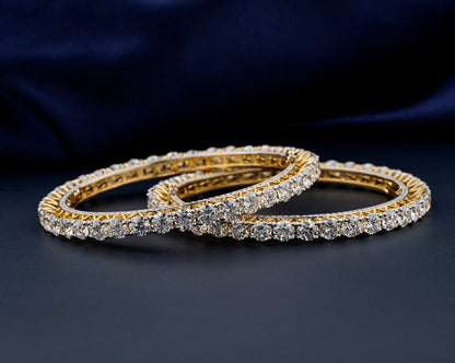 Bracelets with yellow metal bands, round stone tennis design, overlapping pair displayed flat, front angled view against a dark blue fabric background
