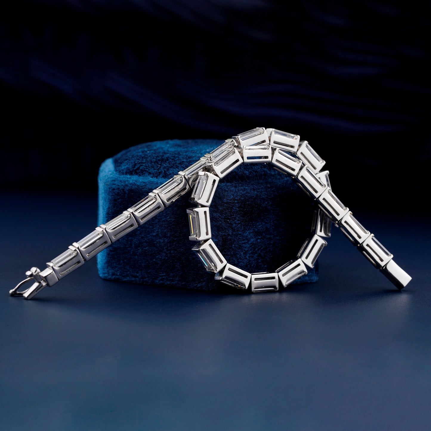 Bracelet with white metal links, rectangular step-cut stones in tennis bracelet design, draped over a blue velvet display, angled front view
