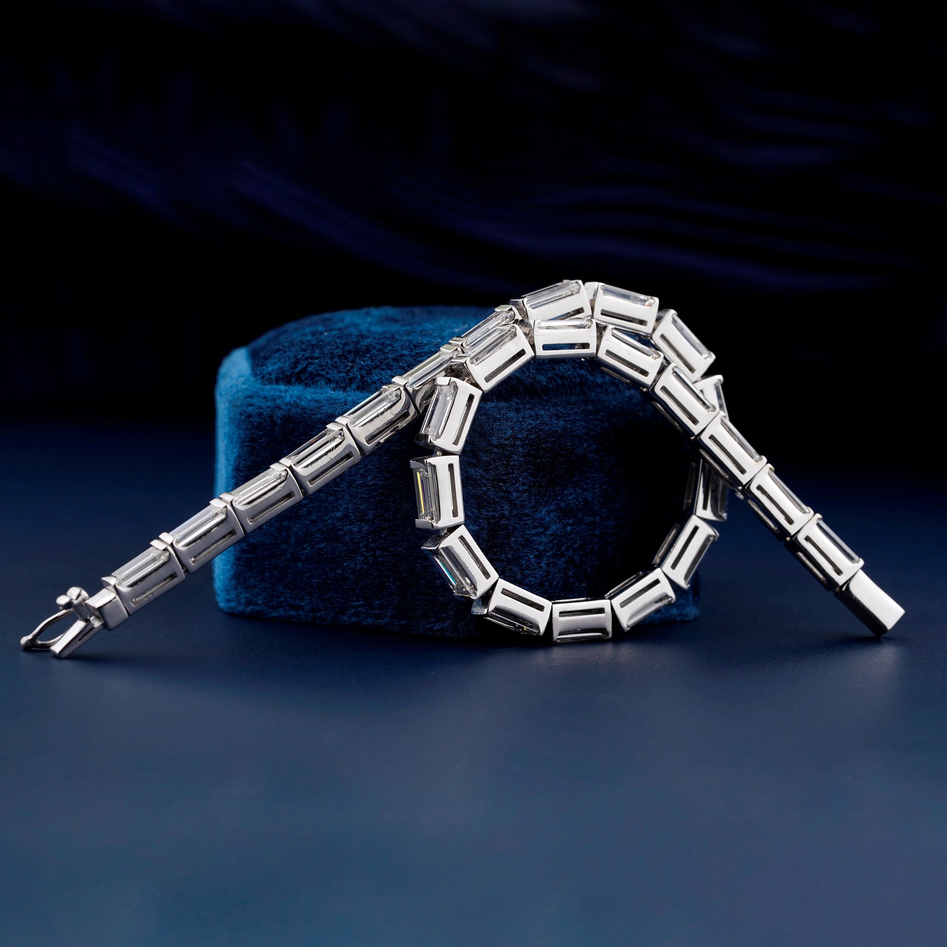 Bracelet with white metal links, rectangular step-cut stones in tennis bracelet design, draped over a blue velvet display, angled front view
