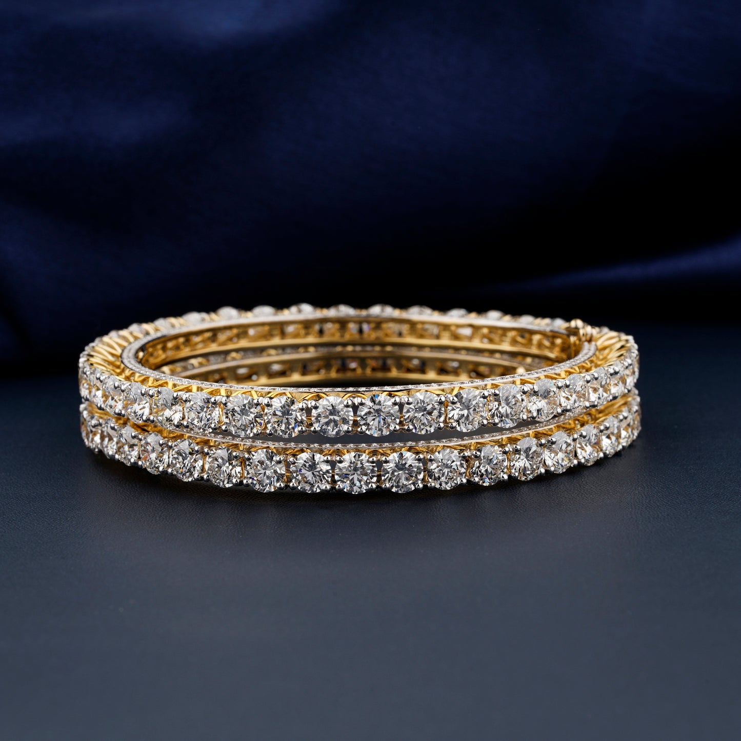 Bracelets with yellow metal bands, round stone tennis design, pair stacked horizontally, front view against a dark blue fabric background
