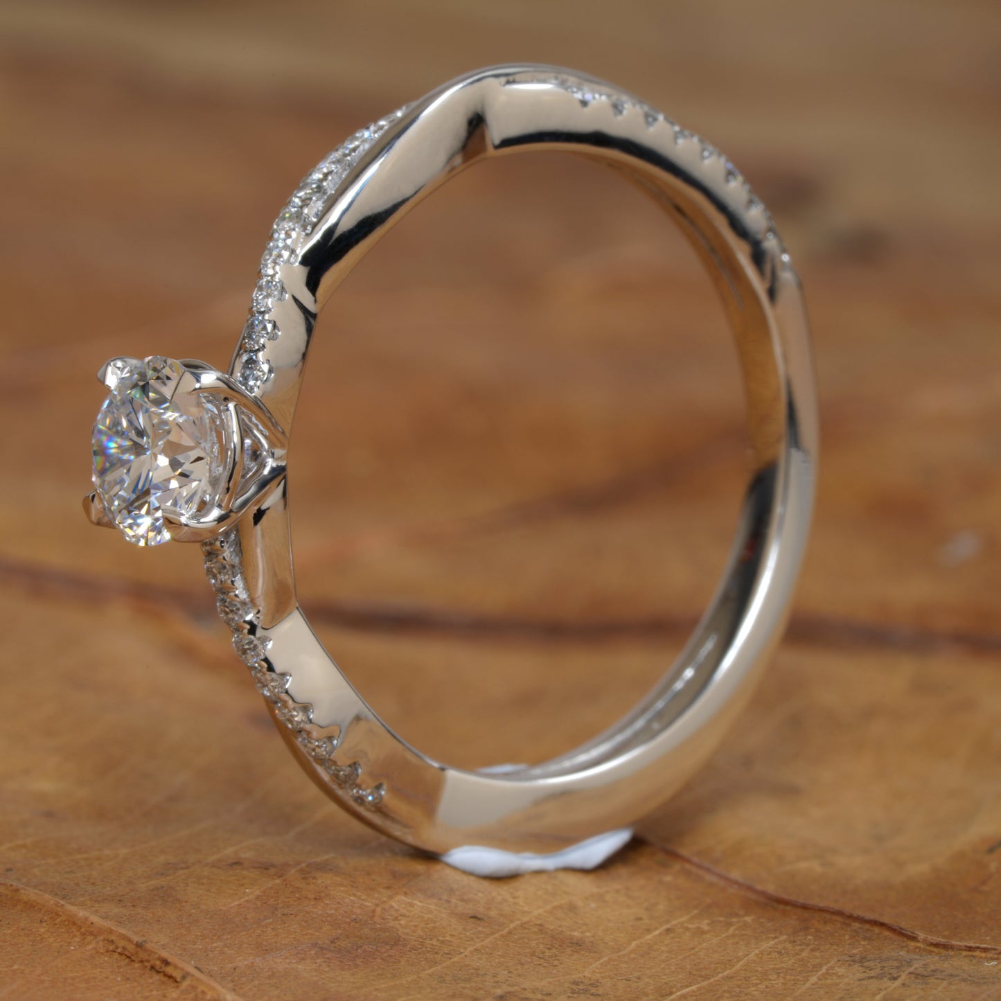 Ring with round clear solitaire, white metal twisted band, pavé-set shoulders, side profile on wooden surface
