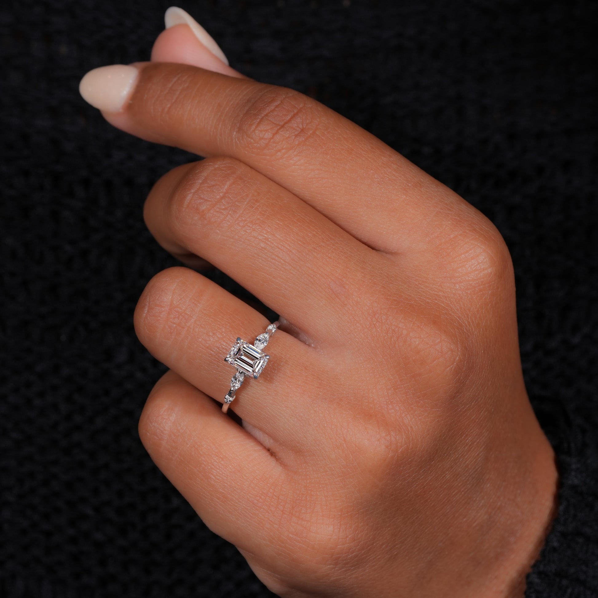 Engagement ring with white metal band, emerald-cut clear center stone, marquise side stones and round accents, worn on finger at angled view
