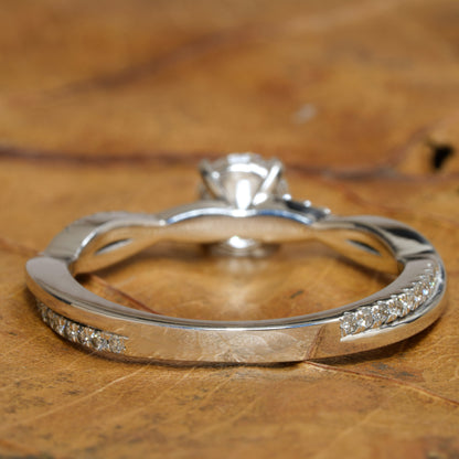 Ring with twisted white metal band, pavé-set shoulders, round solitaire setting visible from the back, rear view on wooden surface

