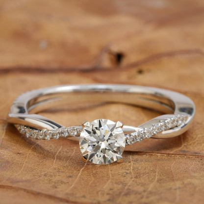 Ring with white metal band, round clear center stone, twisted split shank and pavé-set shoulders, front view on a wooden surface
