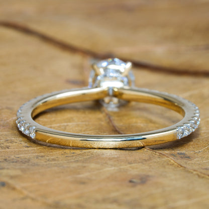 Engagement ring with round clear center stone, white metal band with partial pavé shoulders, rear view on wooden surface
