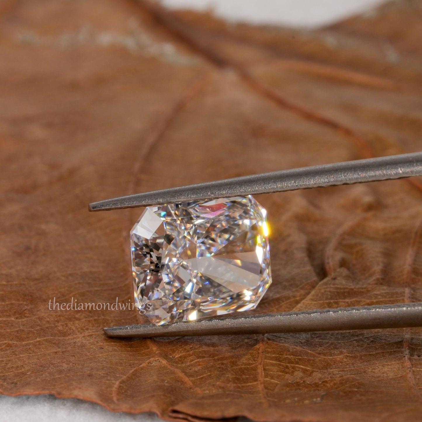 Loose cushion-cut diamond, clear faceted gemstone held with tweezers, angled view showing pavilion facets with partial watermark visible
