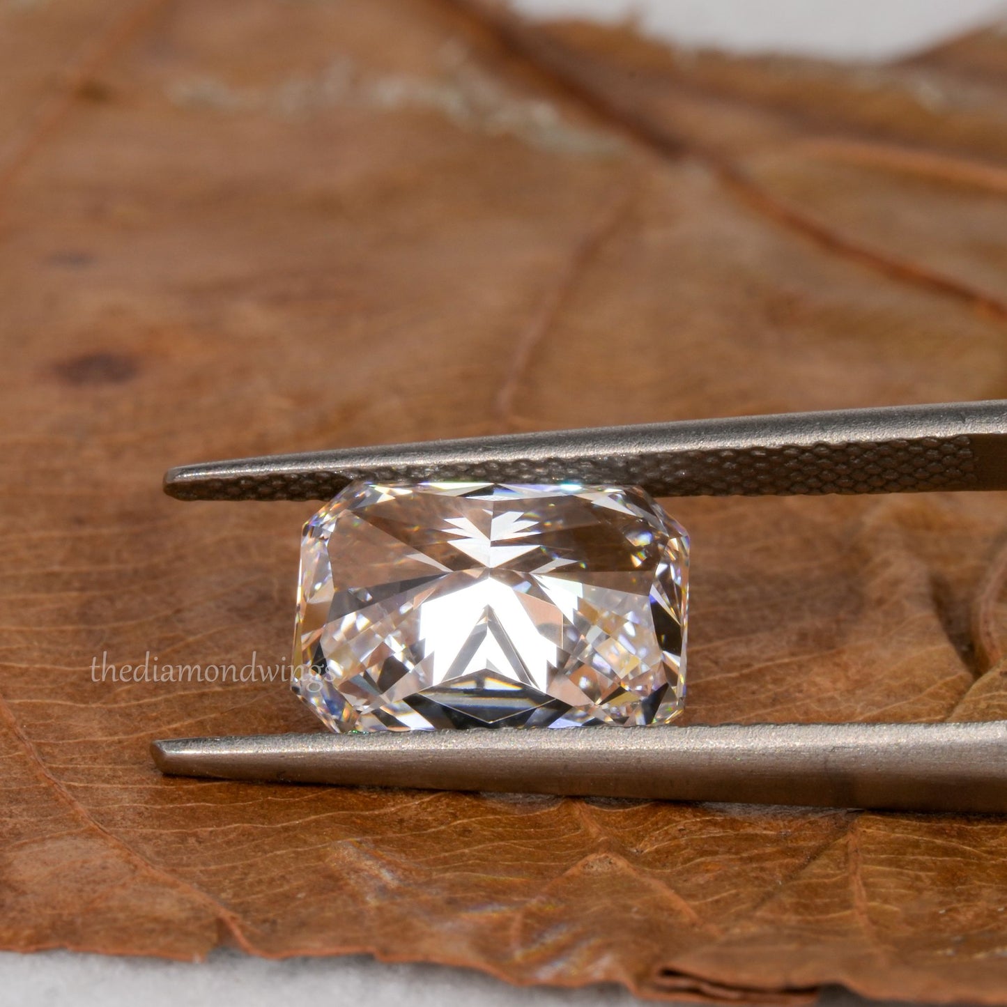 Loose cushion-cut diamond, clear faceted gemstone held with tweezers, pavilion view showing underside facets with partial watermark visible
