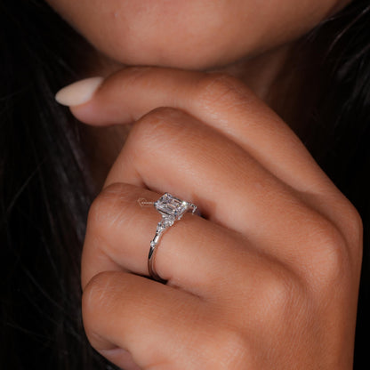 Engagement ring with white metal band, emerald-cut clear center stone, marquise side stones and round accents, worn on finger at angled view
