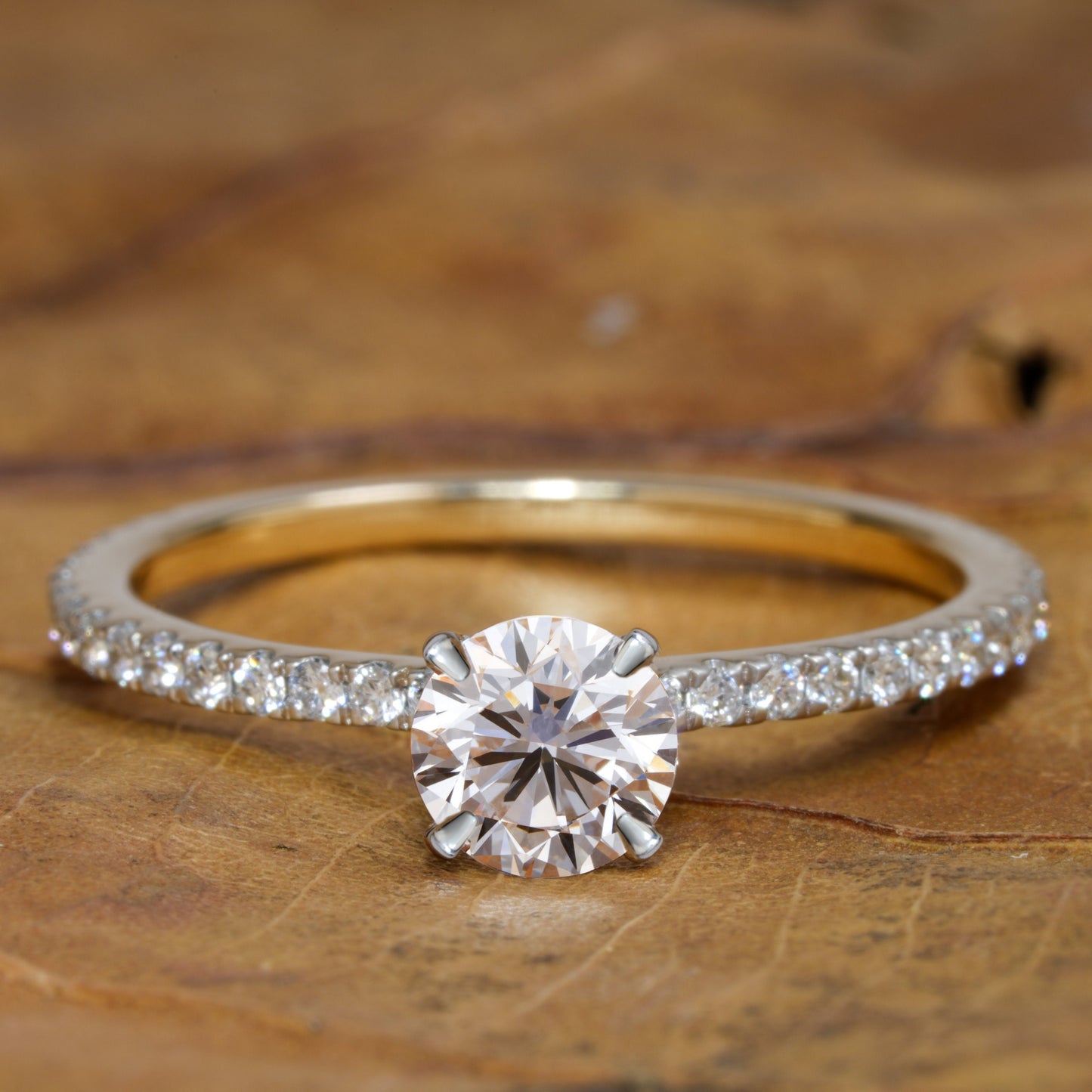Engagement ring with round clear center stone, white metal pavé band, four-prong solitaire setting, front view on wooden surface
