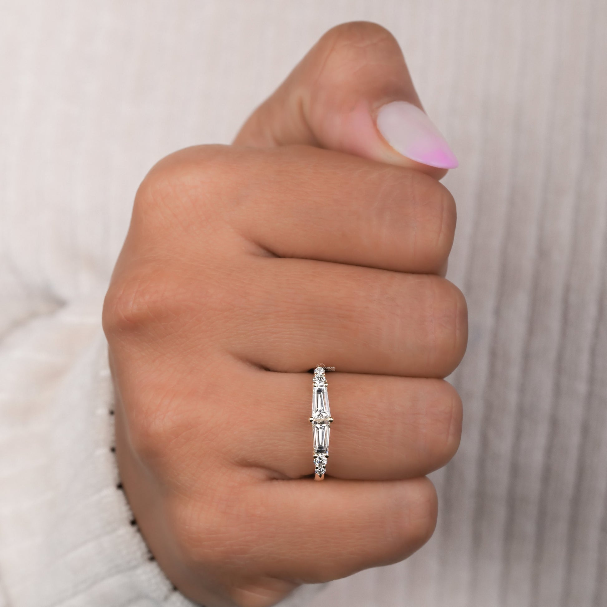 Engagement ring with yellow gold band, vertical baguette-cut clear center gemstone and round side stones, worn on finger, front view
