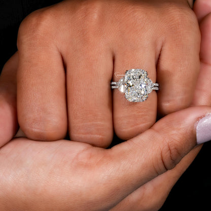 Engagement ring with white metal split shank, elongated cushion-cut clear center stone, oval side stones, worn on finger, front view
