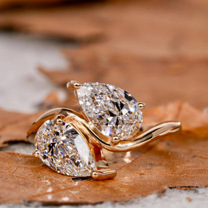 Engagement ring with yellow metal bypass band, two pear-shaped clear gemstones, crossover design, angled close-up view

