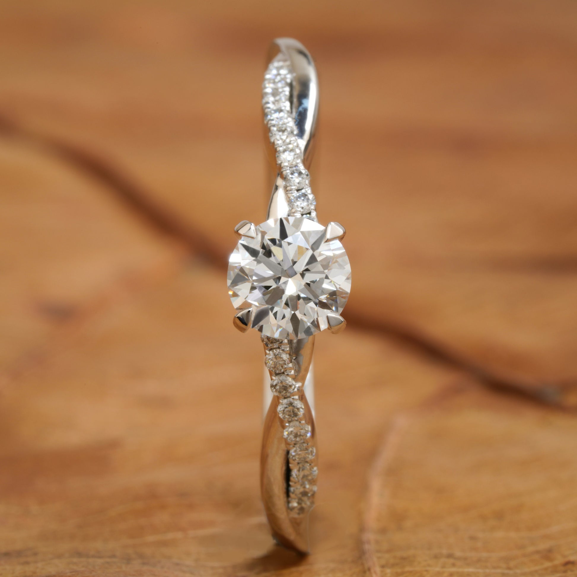 Ring with round clear center stone, twisted white metal band with pavé-set shoulders, front view standing upright on a wooden surface

