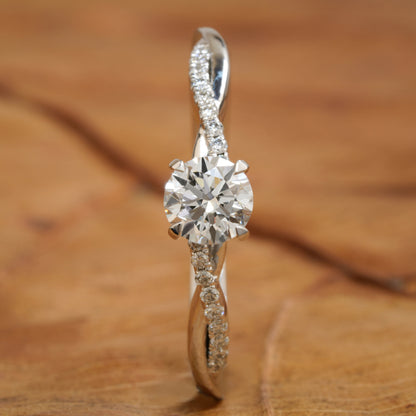 Ring with round clear center stone, twisted white metal band with pavé-set shoulders, front view standing upright on a wooden surface

