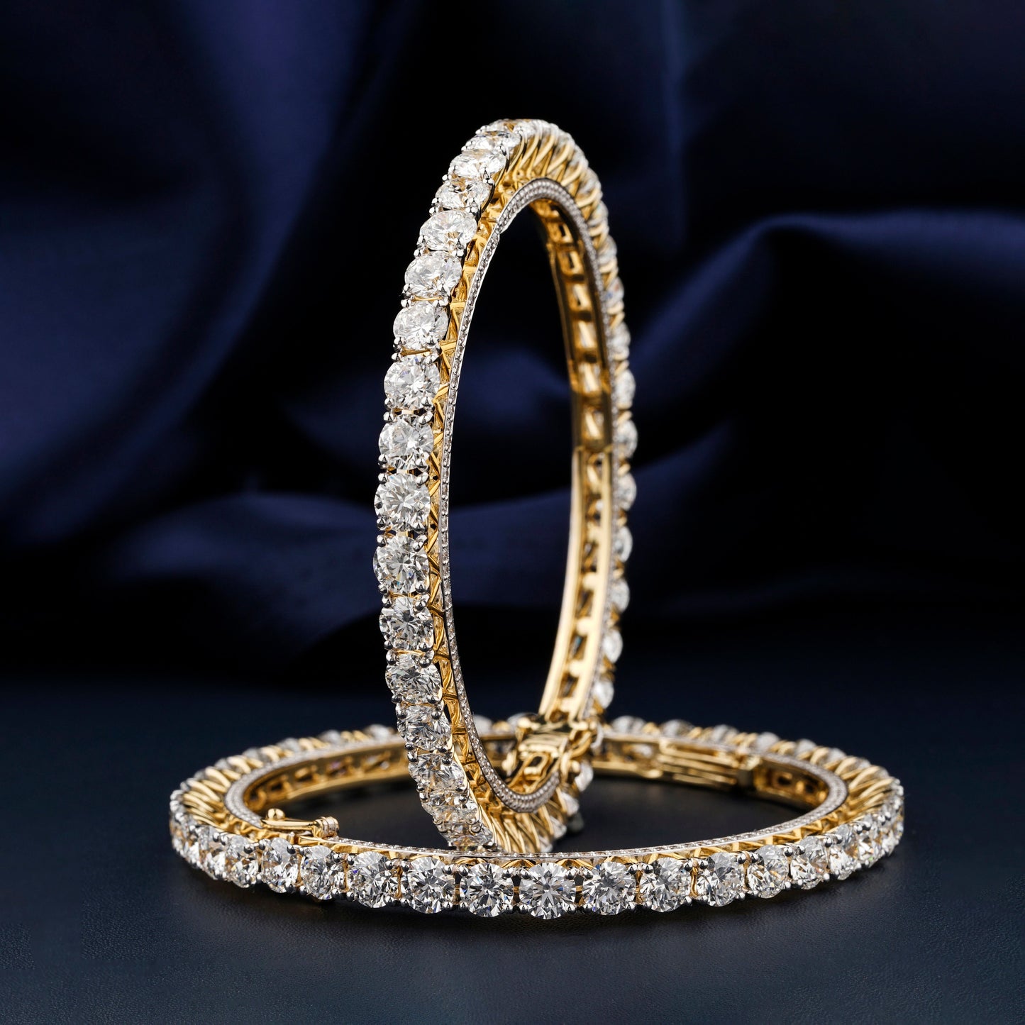 Bracelets with yellow metal bands, round stone tennis design, pair displayed upright and flat, front angled view against a dark blue fabric background
