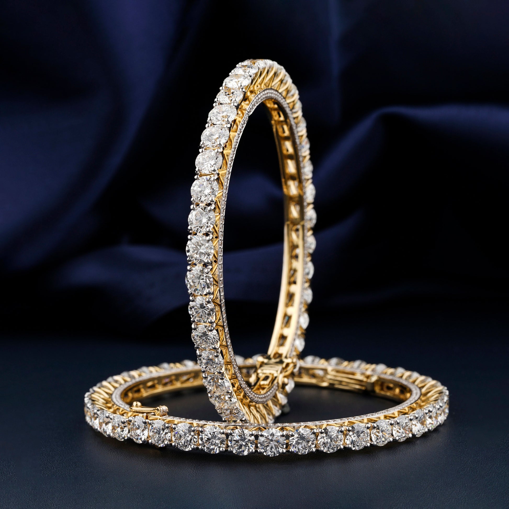 Bracelets with yellow metal bands, round stone tennis design, pair displayed upright and flat, front angled view against a dark blue fabric background
