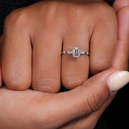 Engagement ring with white metal band, emerald-cut clear center stone, marquise side stones and round accents, worn on finger front view
