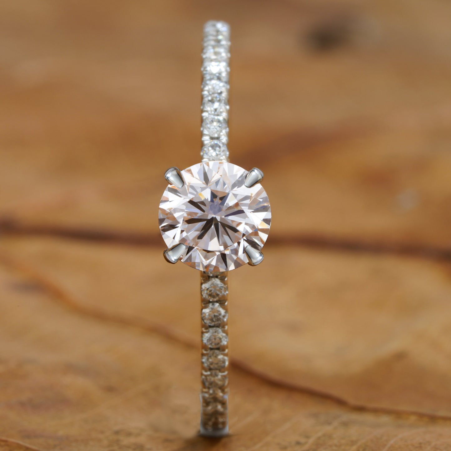 Engagement ring with round clear center stone, white metal pavé band, four-prong solitaire setting, front profile view on wooden surface
