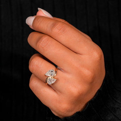 Engagement ring with yellow metal bypass band, two pear-shaped clear gemstones, worn on finger, angled front view

