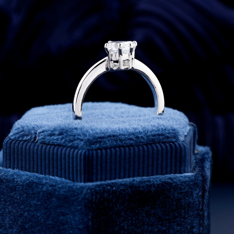 Engagement ring with white metal band, raised prong-set clear center stone, rear profile view on blue velvet ring box
