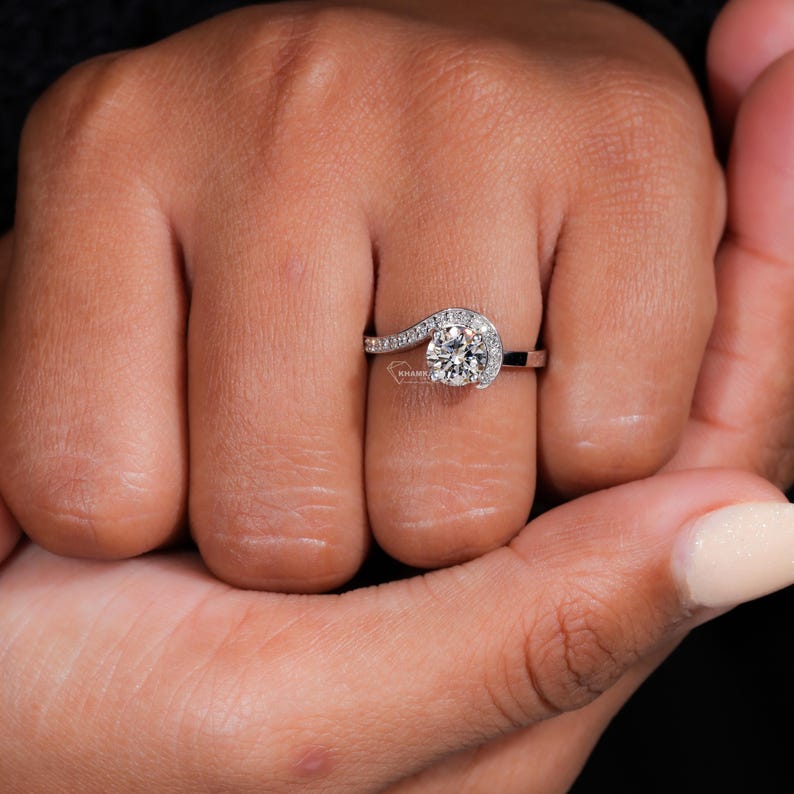 Engagement ring with round center stone, white metal swirl halo design, pavé accent band, worn on hand, front view
