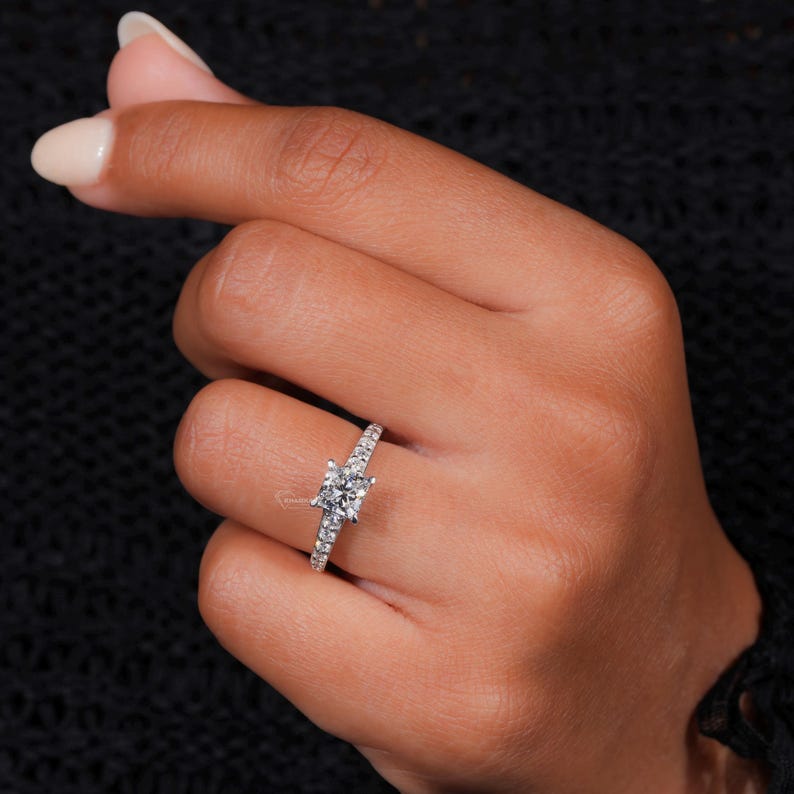 Engagement ring with white metal pavé band, square center stone in four-prong setting, worn on finger, angled front view
