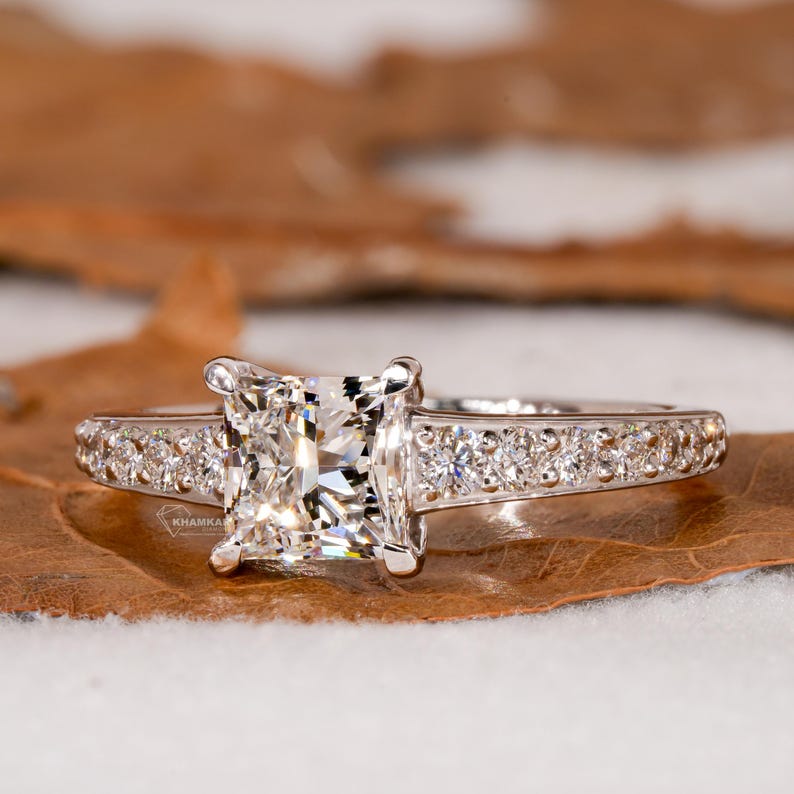 Engagement ring with white metal pavé band, square center stone in four-prong setting, front view on natural surface
