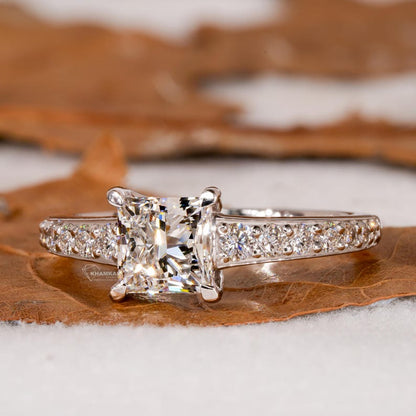 Engagement ring with white metal pavé band, square center stone in four-prong setting, front view on natural surface
