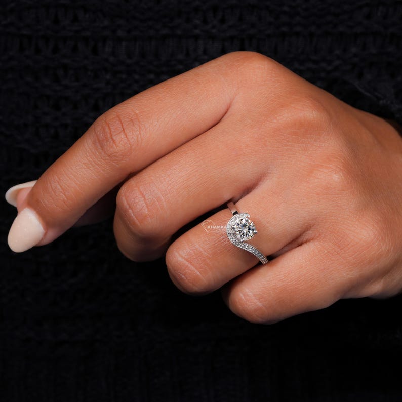 Engagement ring with round center stone, white metal swirl halo design, pavé accent band, worn on hand, angled front view
