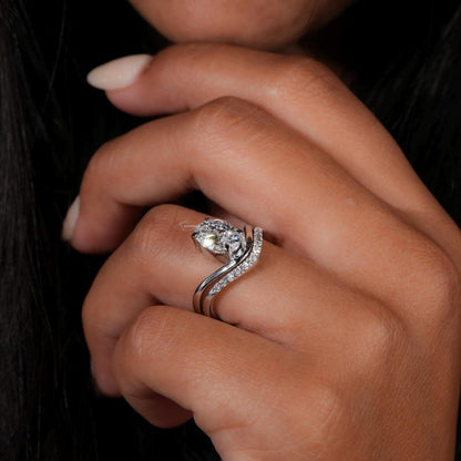 Engagement ring with pear cut clear center stone, silver tone split bypass band with pavé accents, worn on hand, close-up side view