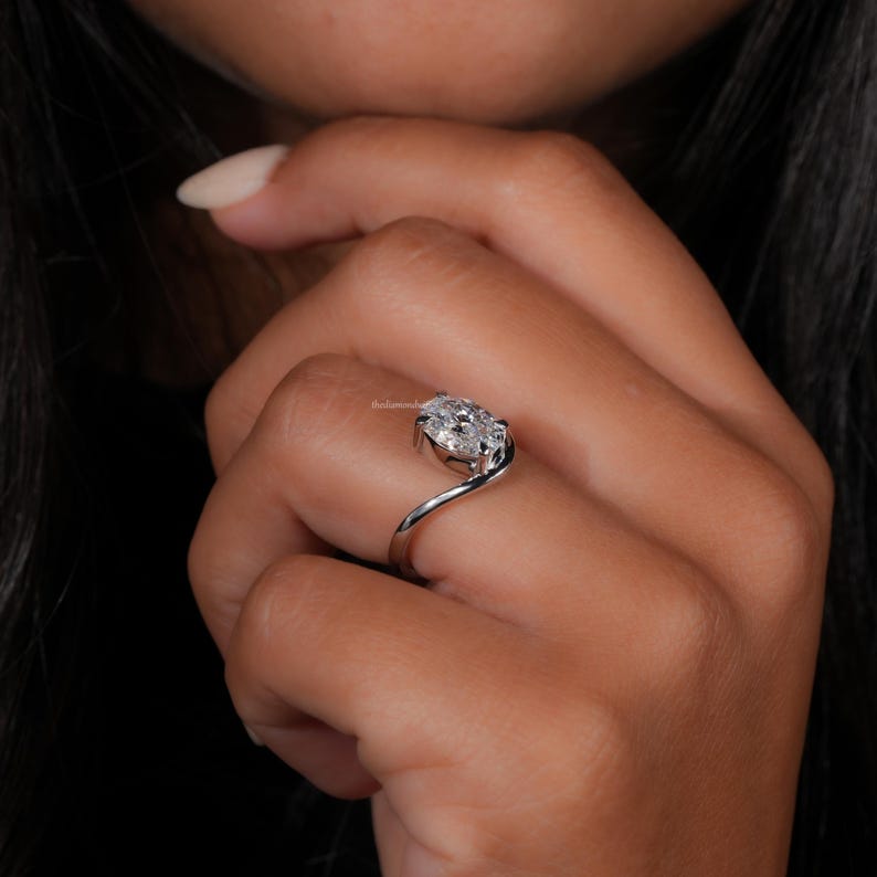 Engagement ring with oval cut clear center stone, silver tone curved solitaire band, worn on hand, angled side view