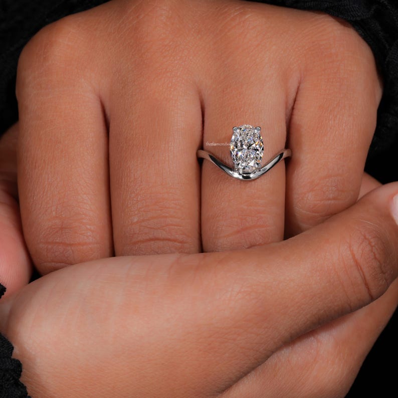 Engagement ring with oval cut clear center stone, silver tone curved solitaire band, worn on hand, front view