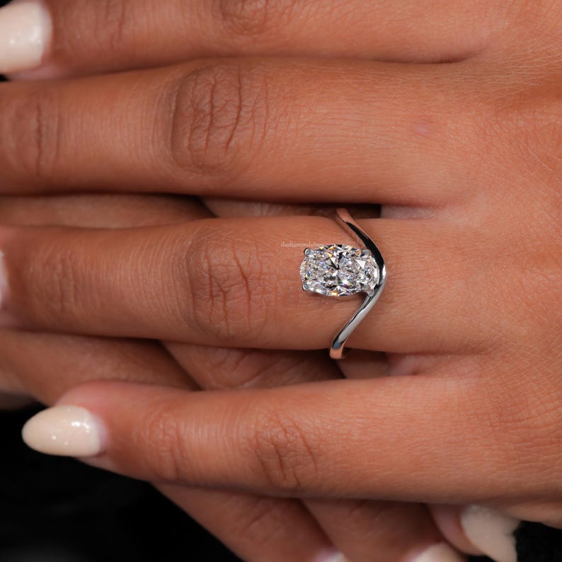 Engagement ring with oval cut clear center stone, silver tone curved solitaire band, worn on hand, angled top view