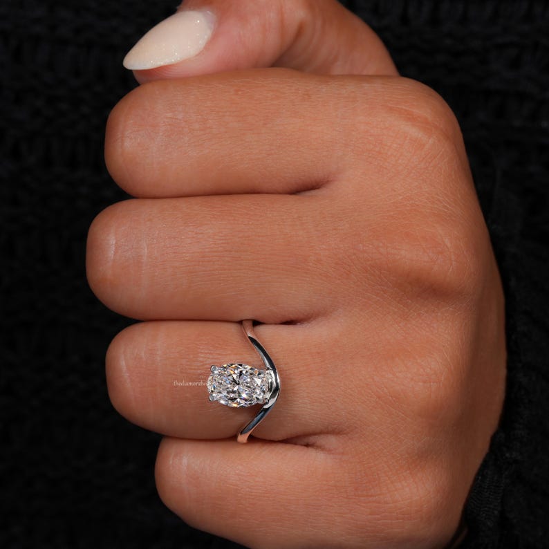 Engagement ring with oval cut clear center stone, silver tone curved solitaire band, worn on hand, close-up front view