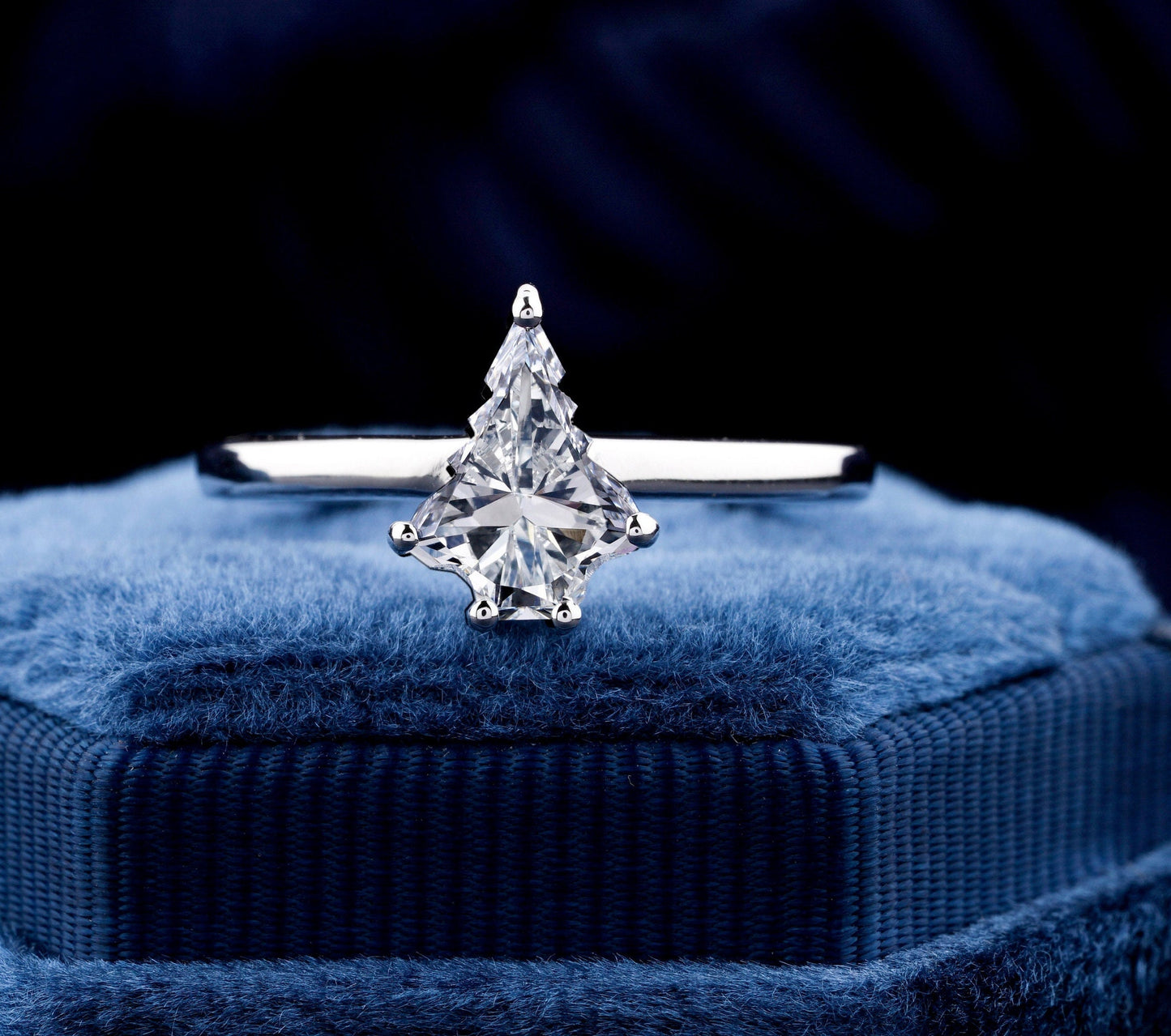 Engagement ring with white metal band, kite-shaped clear center stone in prong setting, front view on blue velvet ring box

