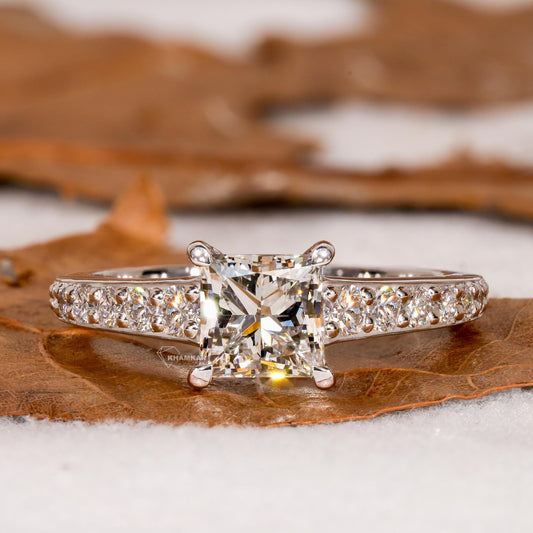 Engagement ring with white metal band, square center stone, pavé band and four-prong setting, front view on natural surface
