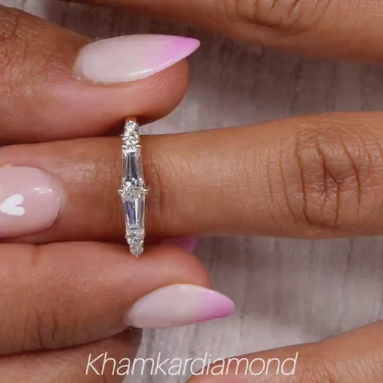 Engagement ring with white metal band, vertical baguette-cut clear center gemstone and round side stones, worn on finger, front view
