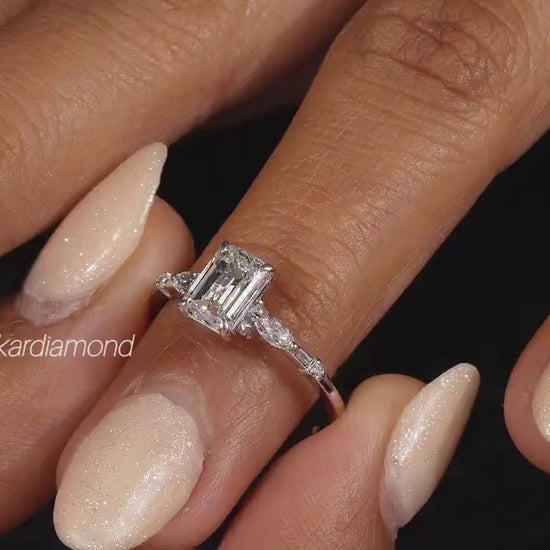 Engagement ring with white metal band, emerald-cut clear center stone, marquise side stones, worn on finger at angled view
